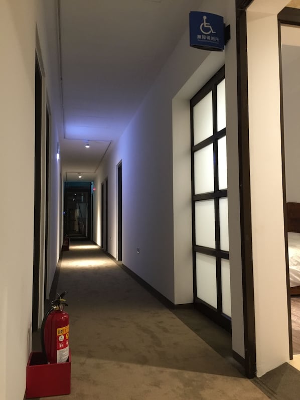 Un pasillo de hotel con un extintor, un letrero de accesibilidad para sillas de ruedas y un ventanal grande a la derecha.