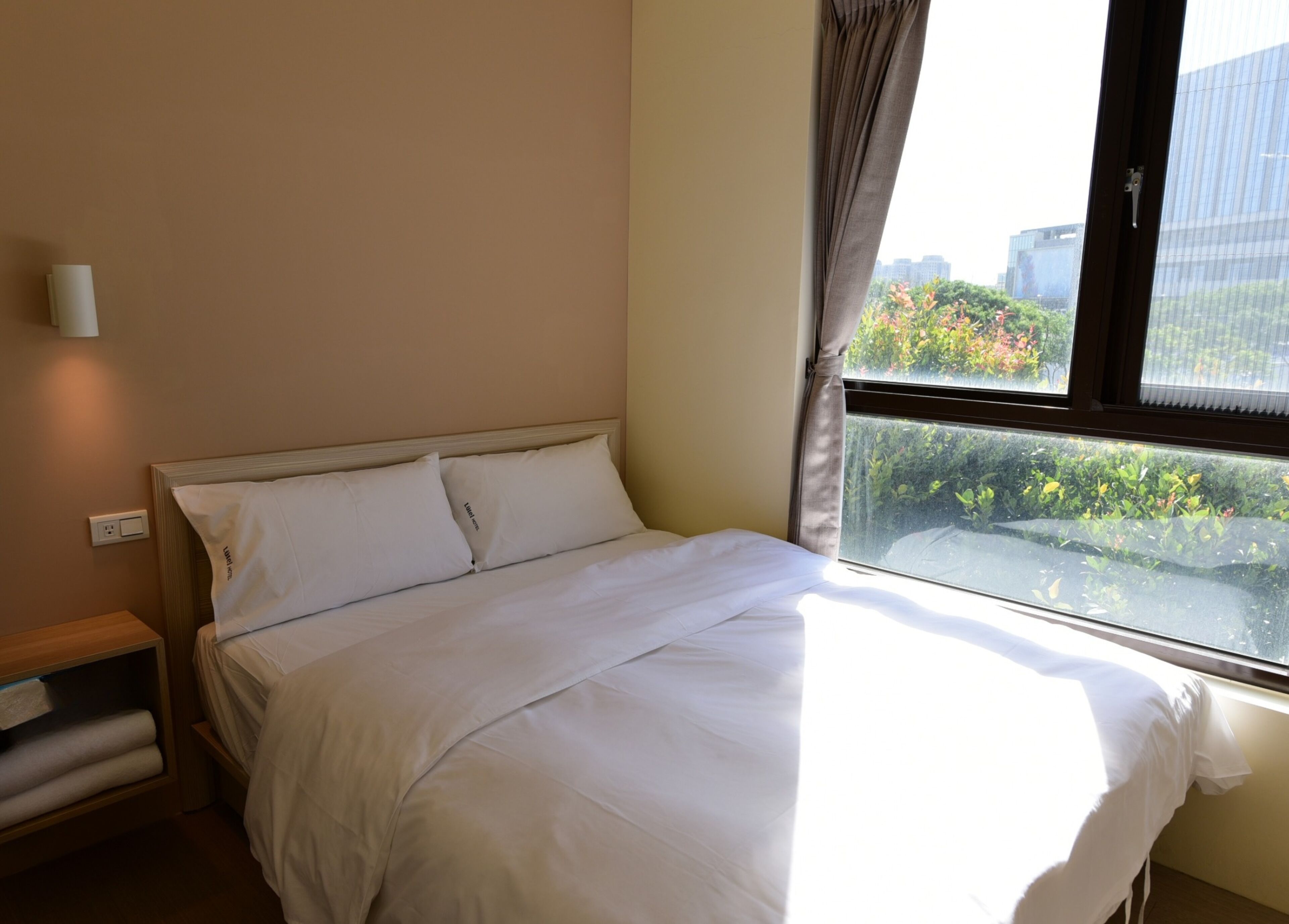 Habitación de hotel con cama, mesita de noche, ventana con cortinas y vistas a zonas verdes y edificios.