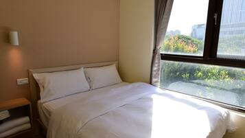 Habitación de hotel con cama, mesita de noche, ventana con cortinas y vistas a zonas verdes y edificios.