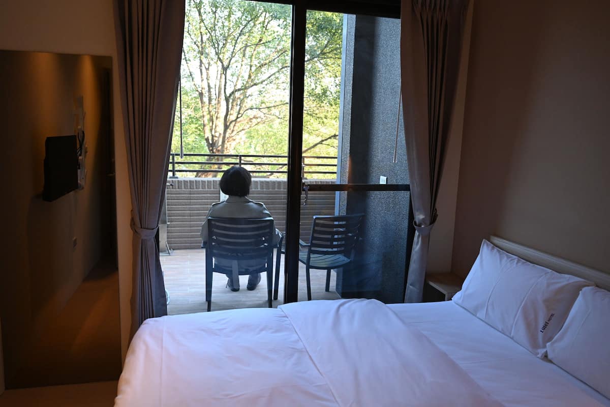 Une chambre d’hôtel avec un lit, une chaise et une vue sur un balcon agrémenté de chaises et d’un arbre.