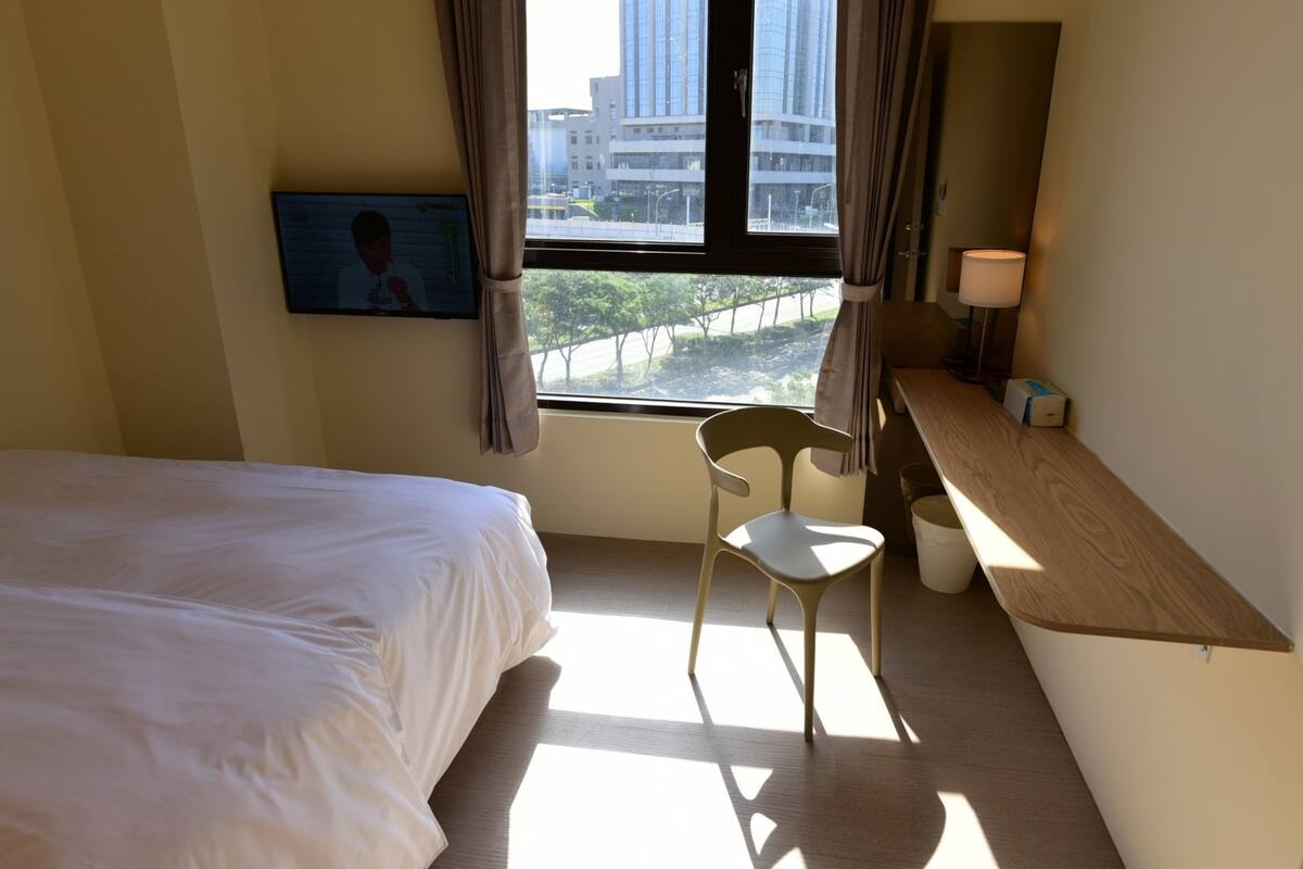 Une chambre d’hôtel avec un lit, une chaise, un bureau, une lampe et une fenêtre avec des rideaux.