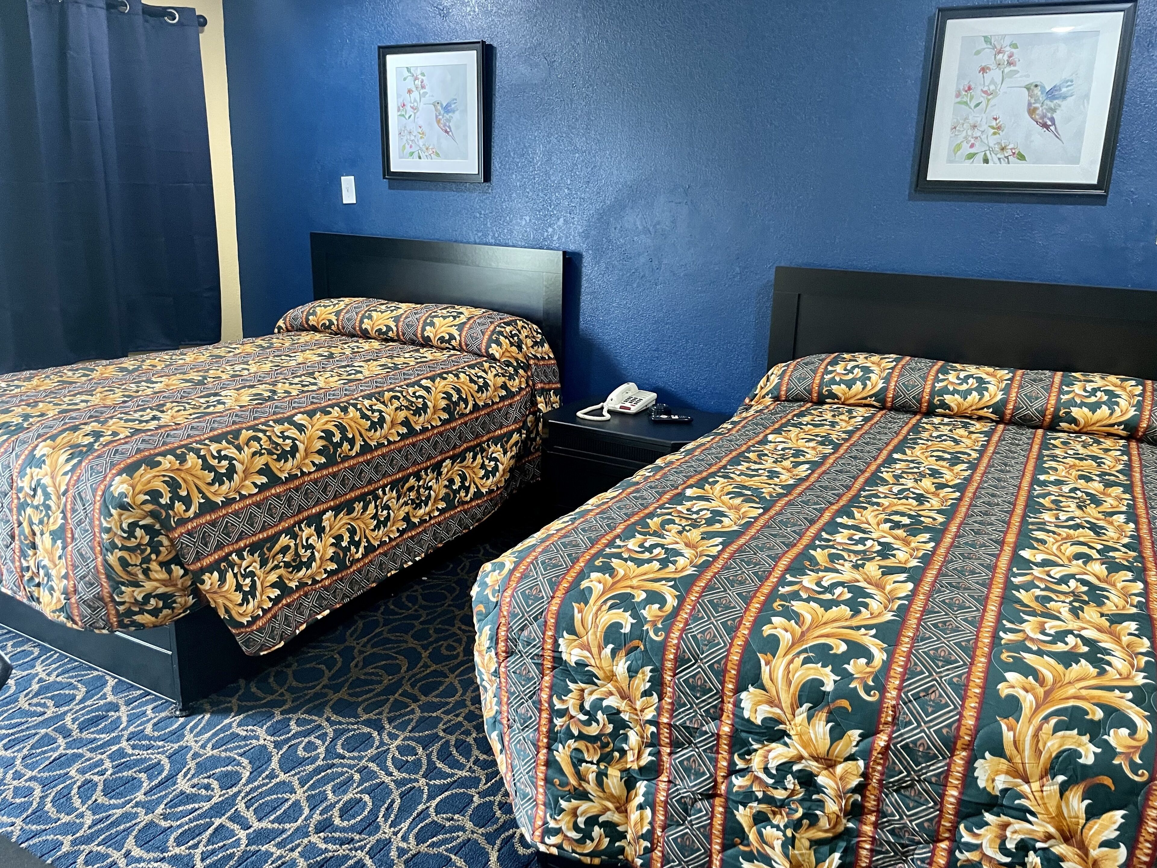 A hotel room with two beds, a dark blue wall, and two framed pictures.