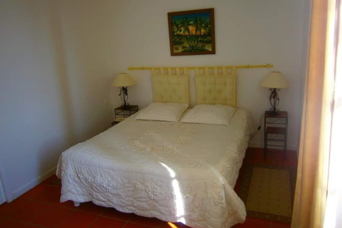 A bedroom with a bed, two bedside tables, a nightstand, a painting on the wall, and a window with curtains.