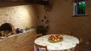 Un comedor rústico con una mesa redonda cubierta por un mantel floral, rodeada de sillas de madera. Se observa un frutero sobre la mesa, y un horno de ladrillo al fondo.