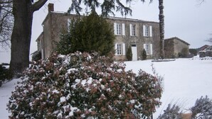 Un edificio de piedra con fachada simétrica, una entrada central y un jardín cubierto de nieve con un arbusto en primer plano.