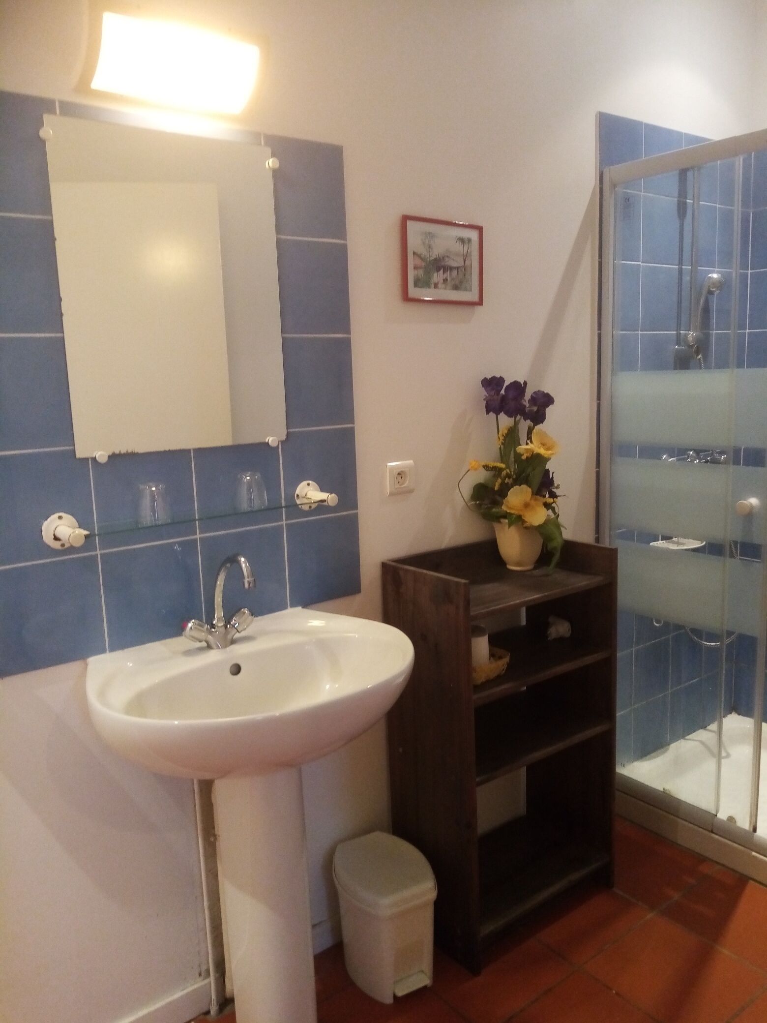 A bathroom with a white sink, blue tiles, a glass shower enclosure, and a small shelf with a vase of flowers.