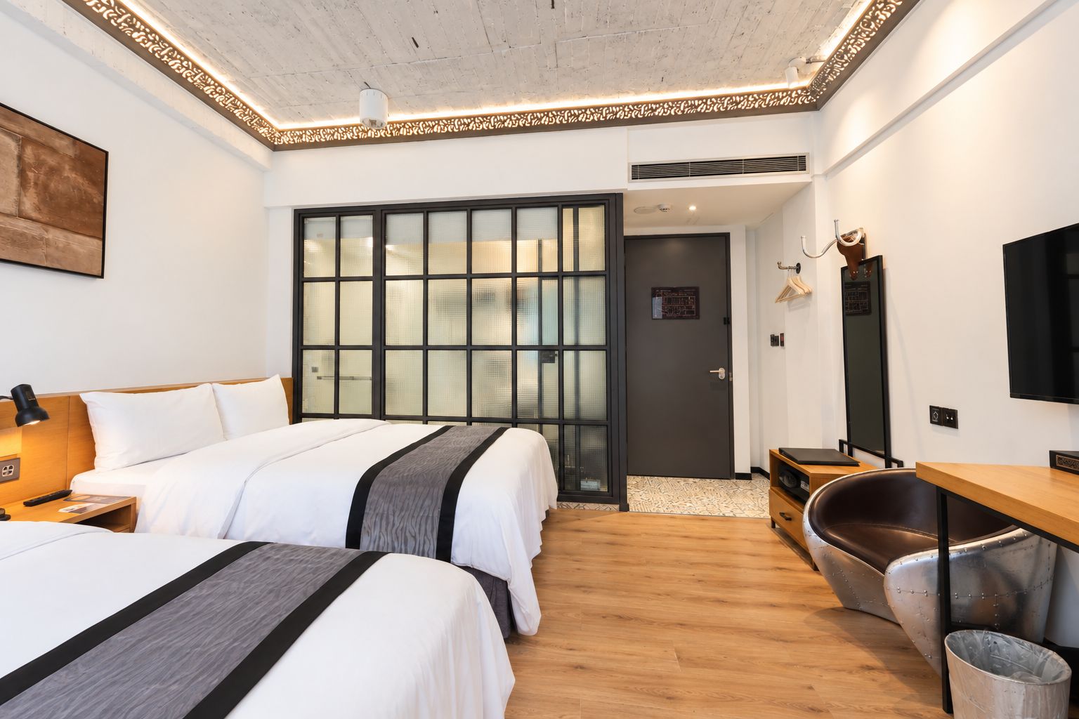 A hotel room with two beds, a desk, a chair, a TV, and a decorative wall panel.