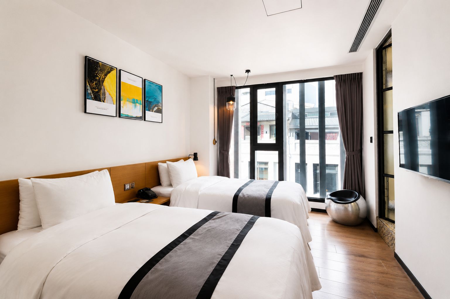 A hotel room with two beds, a large window with curtains, a flat-screen TV, and artwork on the wall.