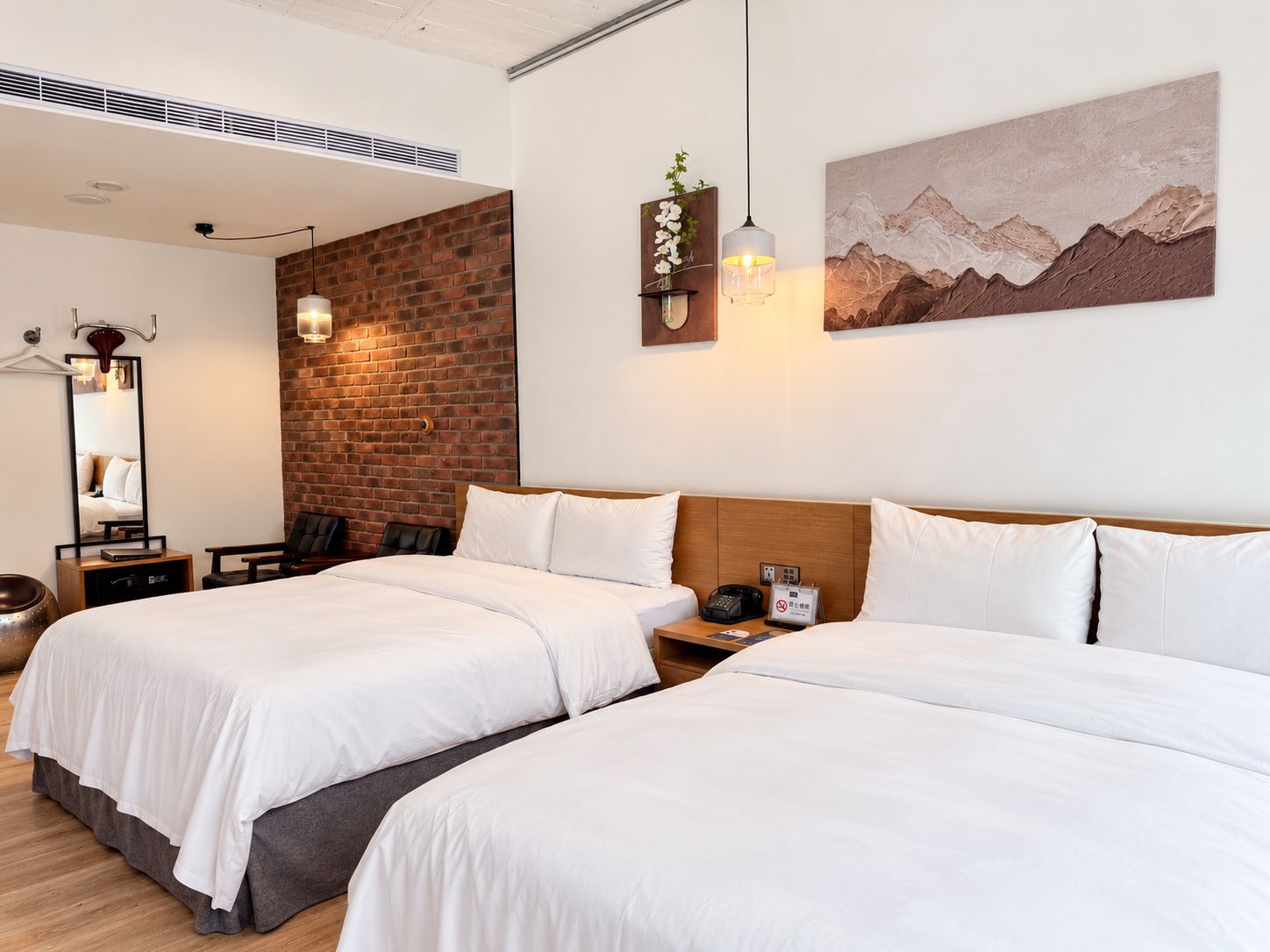 A hotel room with two beds, a brick accent wall, a mountain landscape painting, a hanging light fixture, and a mirror.