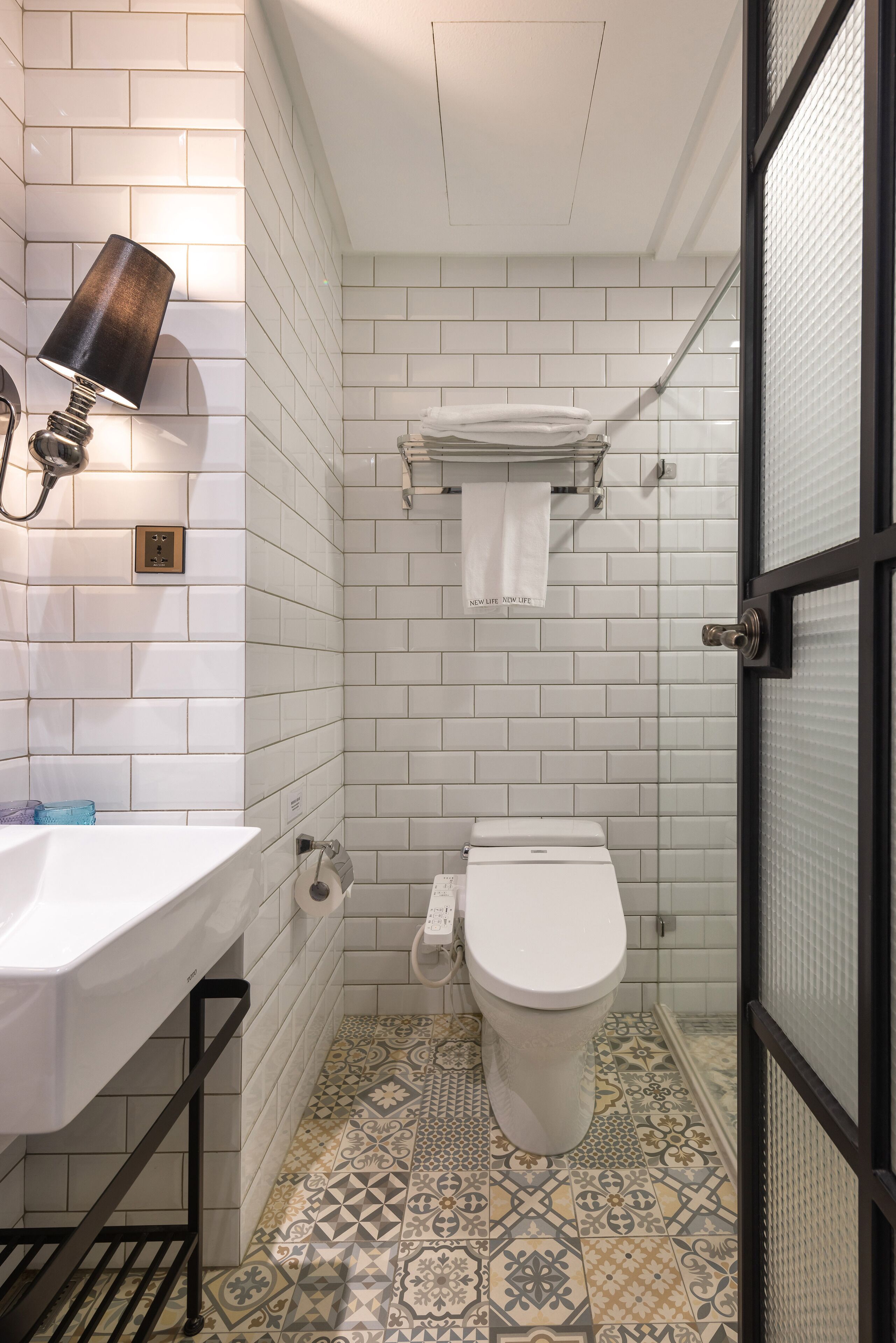 A bathroom with a white toilet, a sink with a black towel holder, patterned floor tiles, and a glass shower door.