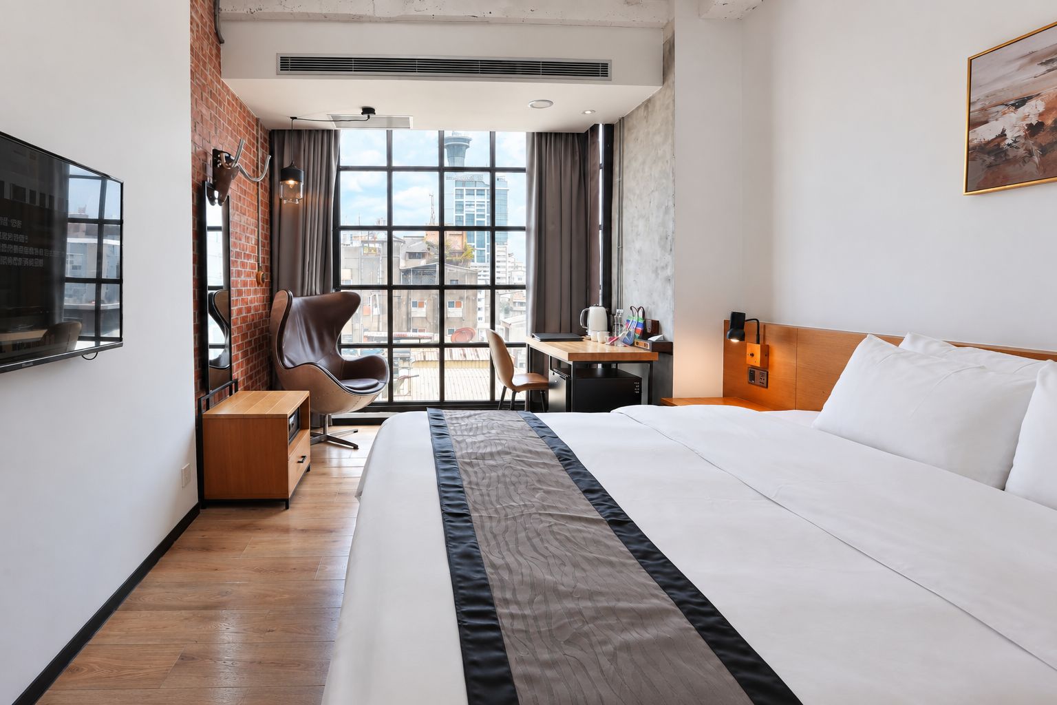 Ett hotellrum med en säng, ett skrivbord, en stol och utsikt över stadsbilden genom stora fönster.