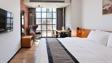 Ett hotellrum med en säng, ett skrivbord, en stol och utsikt över stadsbilden genom stora fönster.
