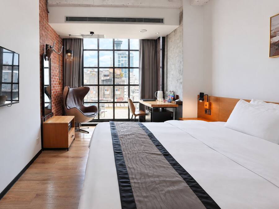 Ett hotellrum med en säng, ett skrivbord, en stol och utsikt över stadsbilden genom stora fönster.