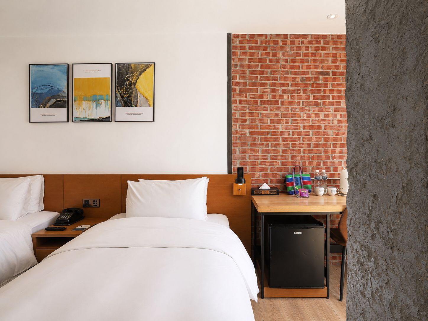 A hotel room with two beds, a desk with a computer, a brick accent wall, and artwork on the wall.