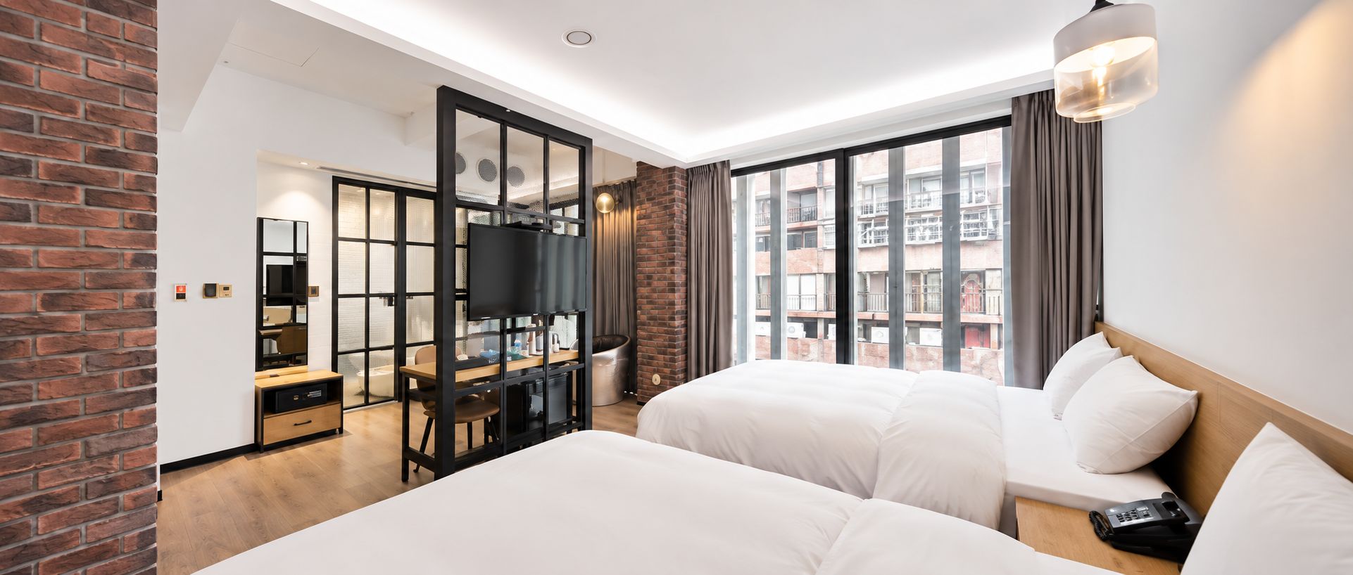 A hotel room with two beds, a desk, a TV, and a large window with a city view.
