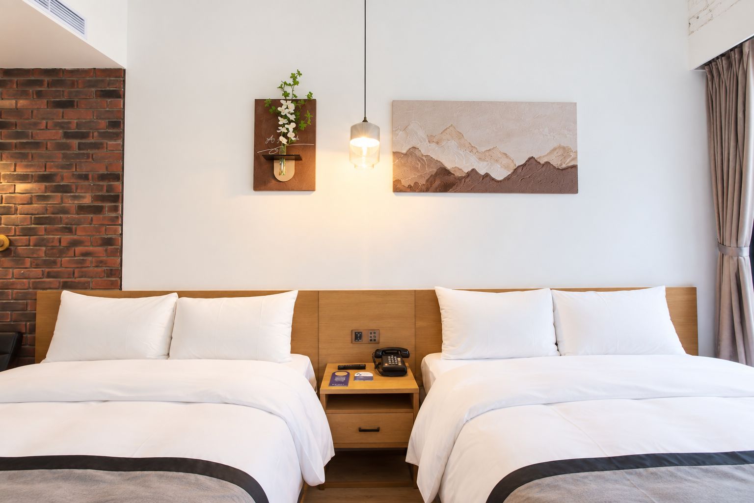 A hotel room with two beds, a brick accent wall, a wall-mounted plant, a painting of mountains, and a bedside table with a phone.
