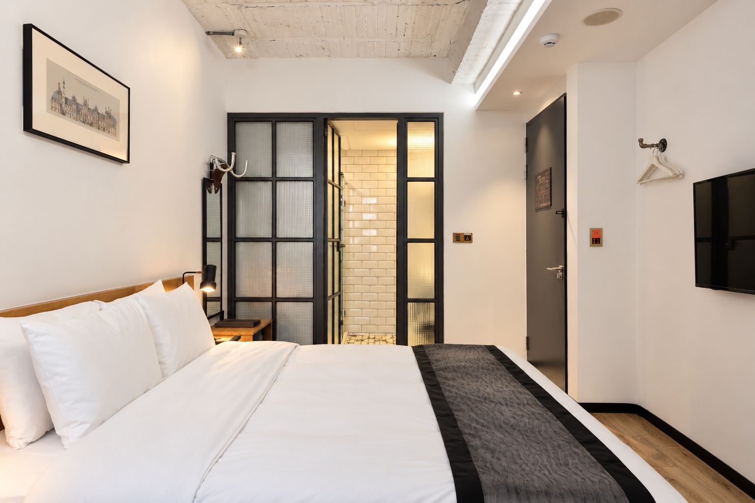 Une chambre d’hôtel avec un lit, un téléviseur fixé au mur, une porte avec un porte-serviettes et un tableau encadré au mur.