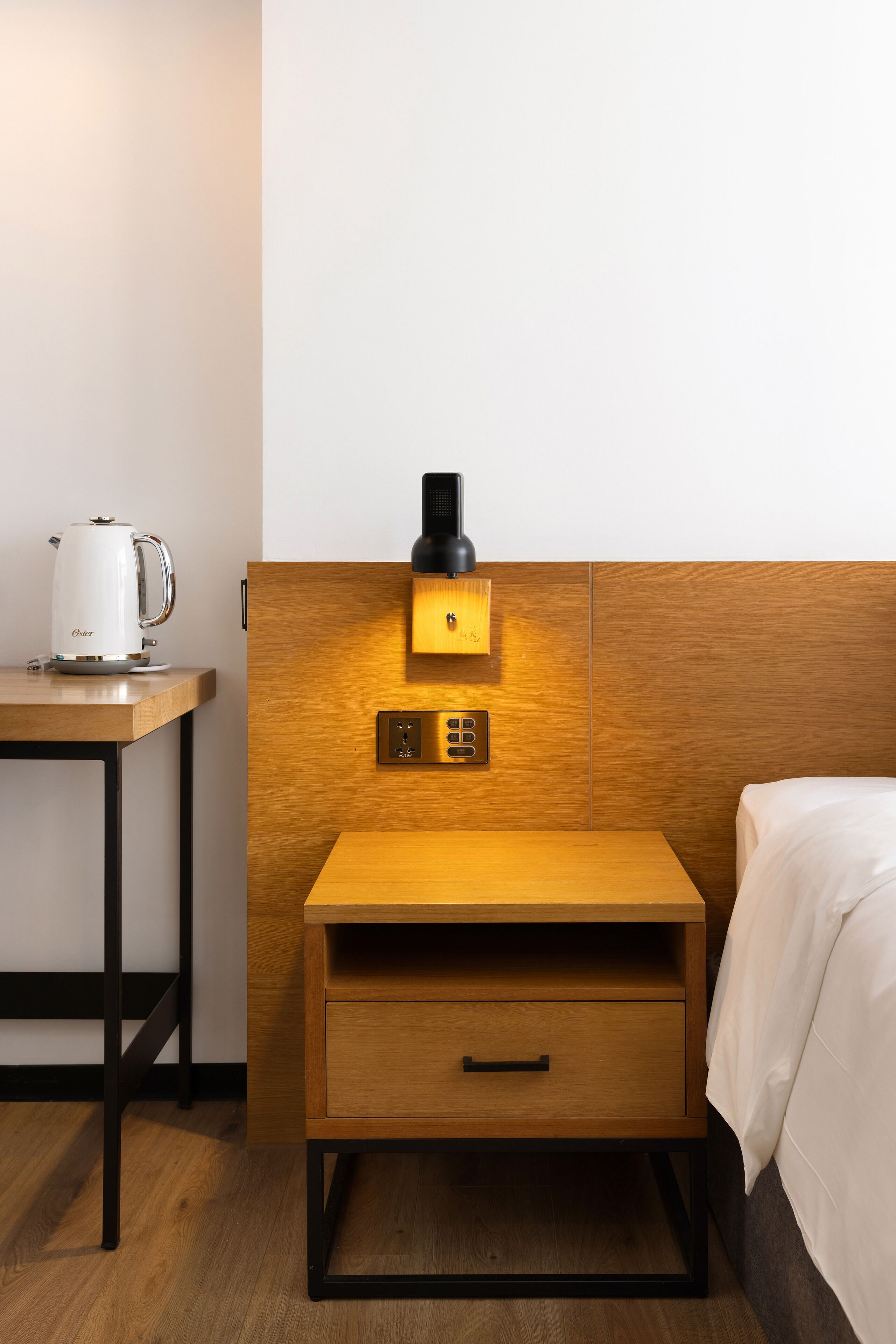 A hotel room with a wooden nightstand, a white kettle, a wall-mounted coffee maker, and a bed with white bedding.