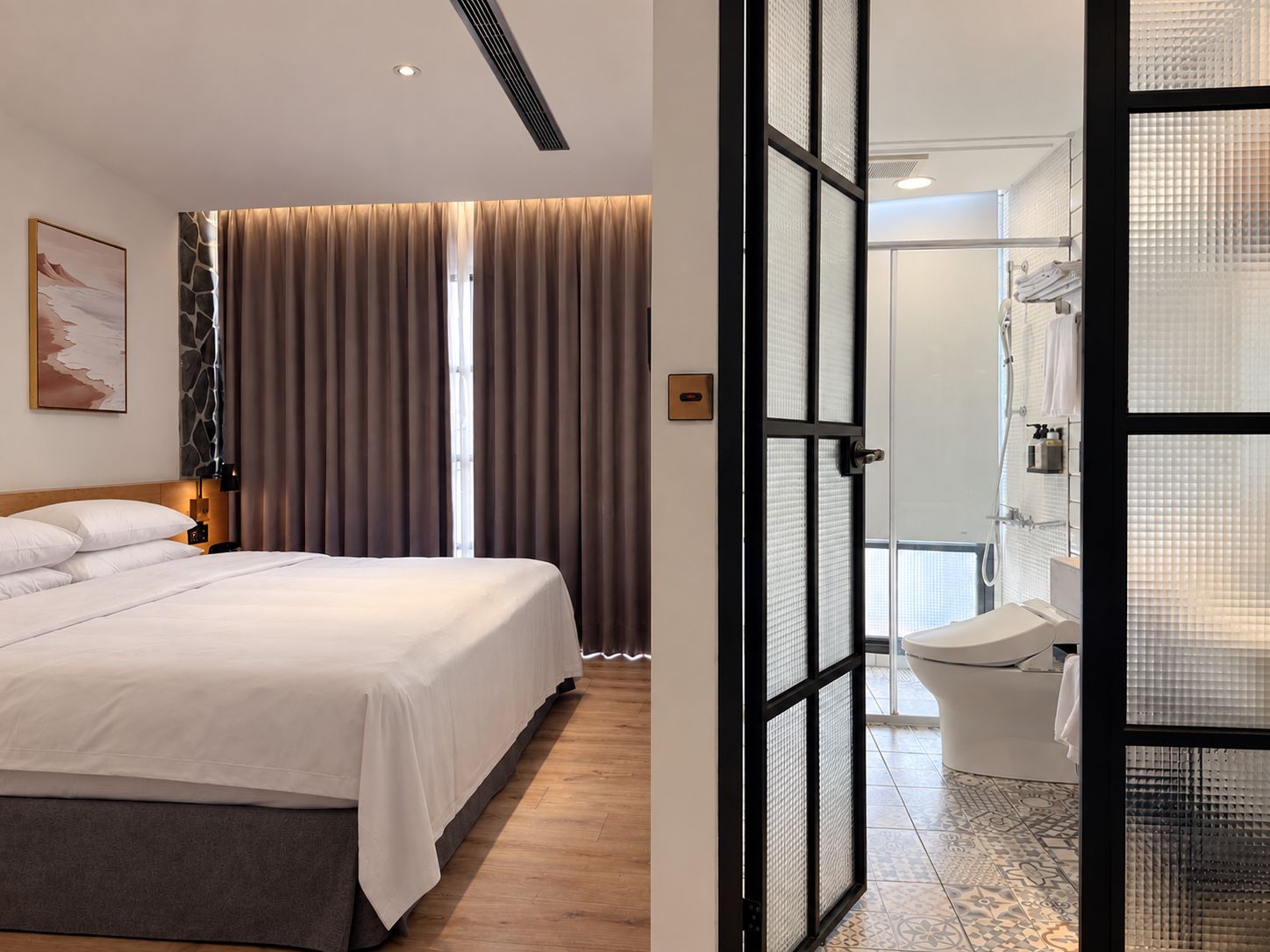 A hotel room with a bed, curtains, a painting, and a bathroom with a toilet and shower.