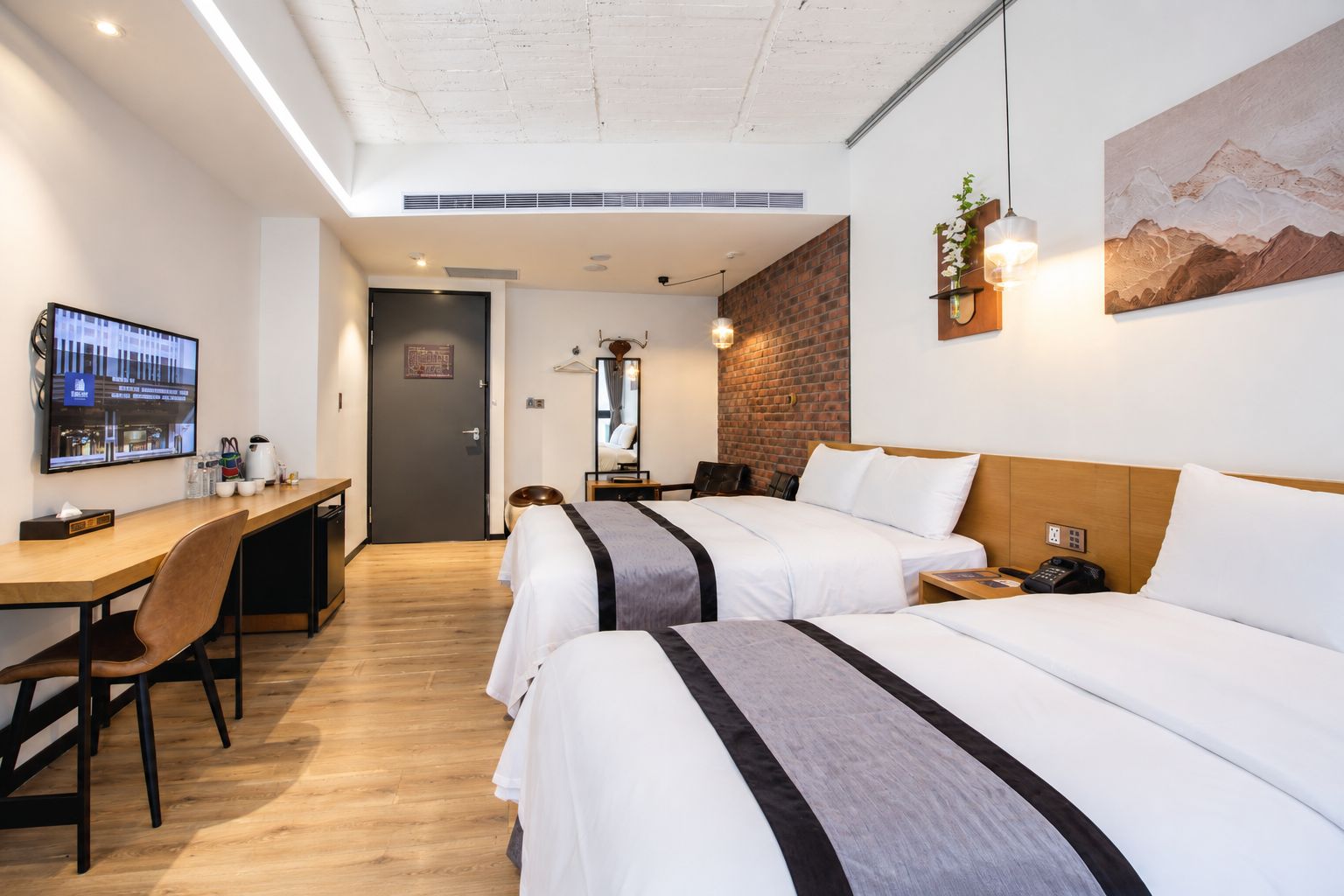 A hotel room with a desk, two beds, a TV, and a mountain painting.