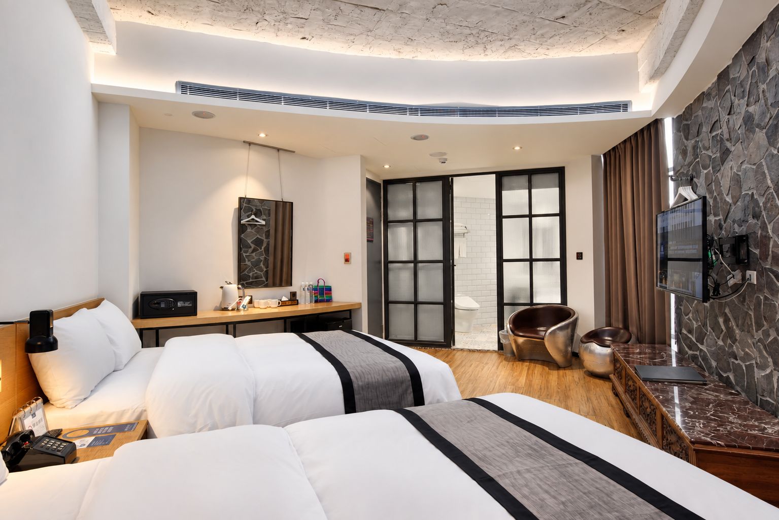 Une chambre d’hôtel avec deux lits, un bureau, une chaise, une télévision et un mur en pierre.