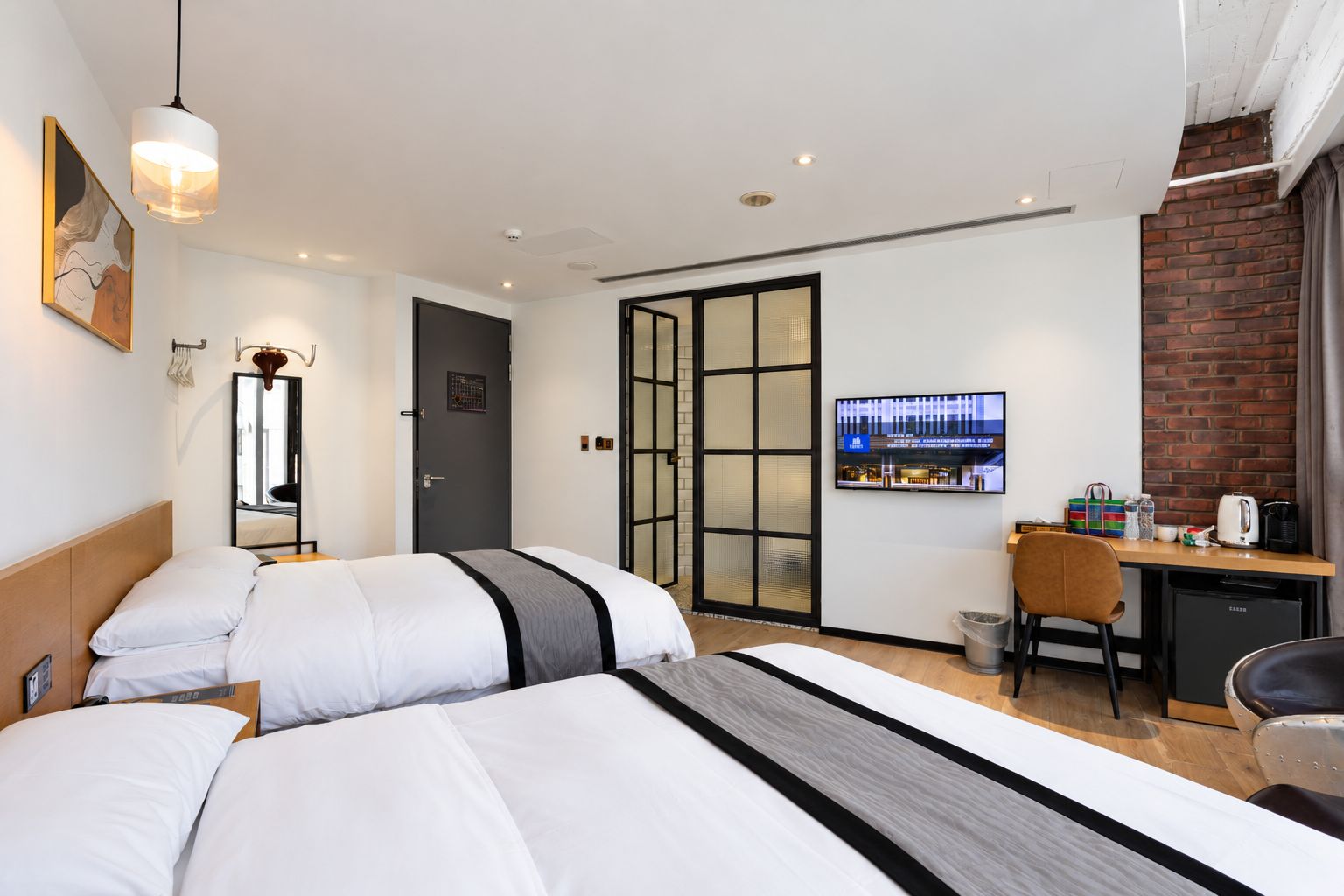 Ein Hotelzimmer mit zwei Betten, einem Schreibtisch, einem Sessel, einem Fernseher und einer Ziegelwand.