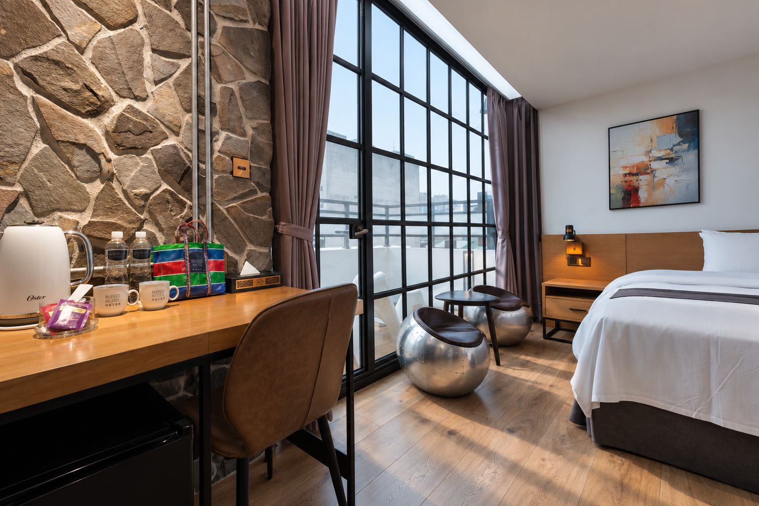 A hotel room with a stone wall, a large window with curtains, a bed, a desk with a coffee maker, and a painting on the wall.