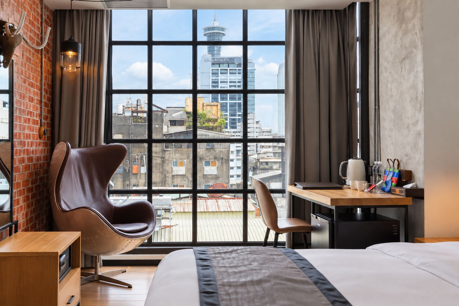 A hotel room with a large window, a bed, a desk with a chair, a lamp, and a brick wall.