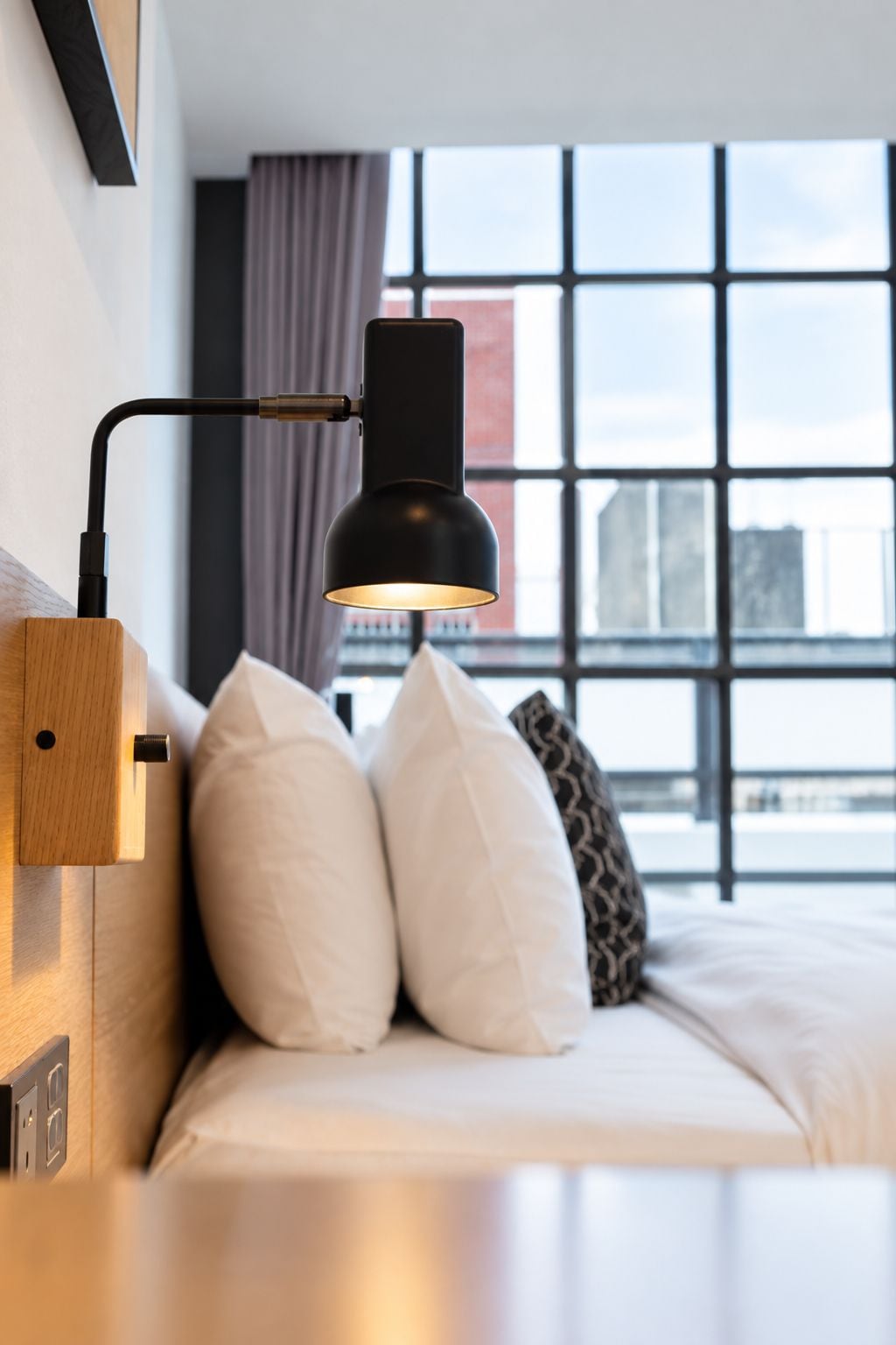A modern bedroom with a bed, a wall-mounted lamp, and a large window with a city view.