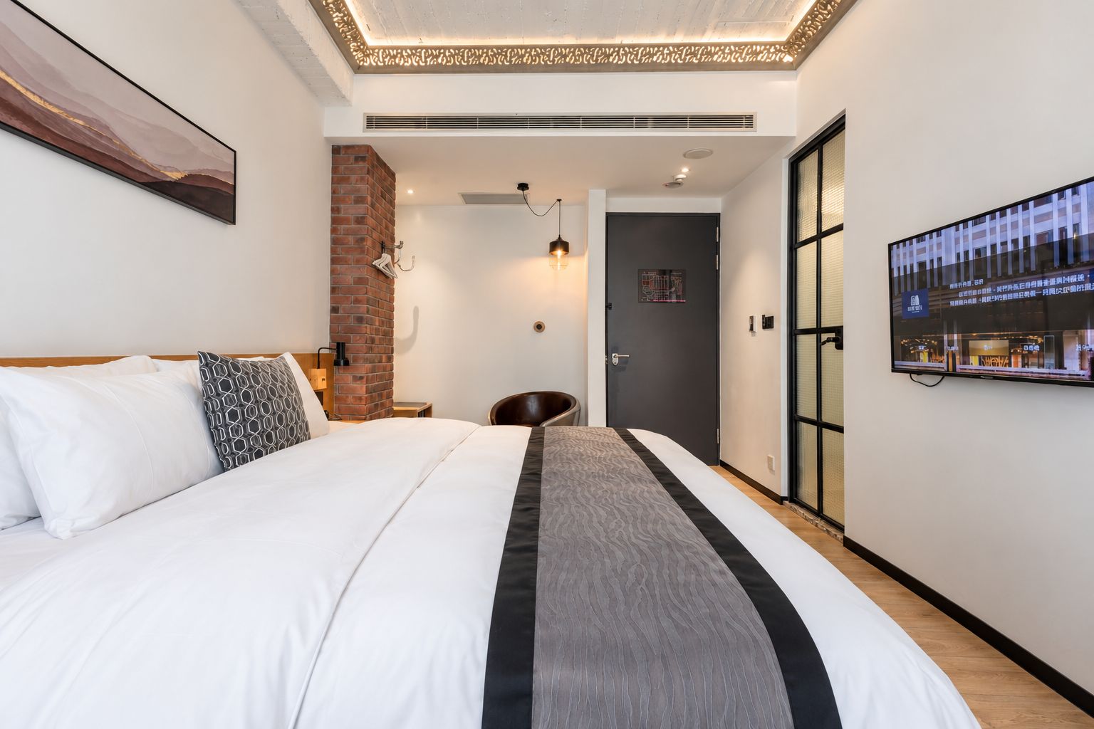 A hotel room with a bed, a brick wall, a desk, a chair, a television, and a door.