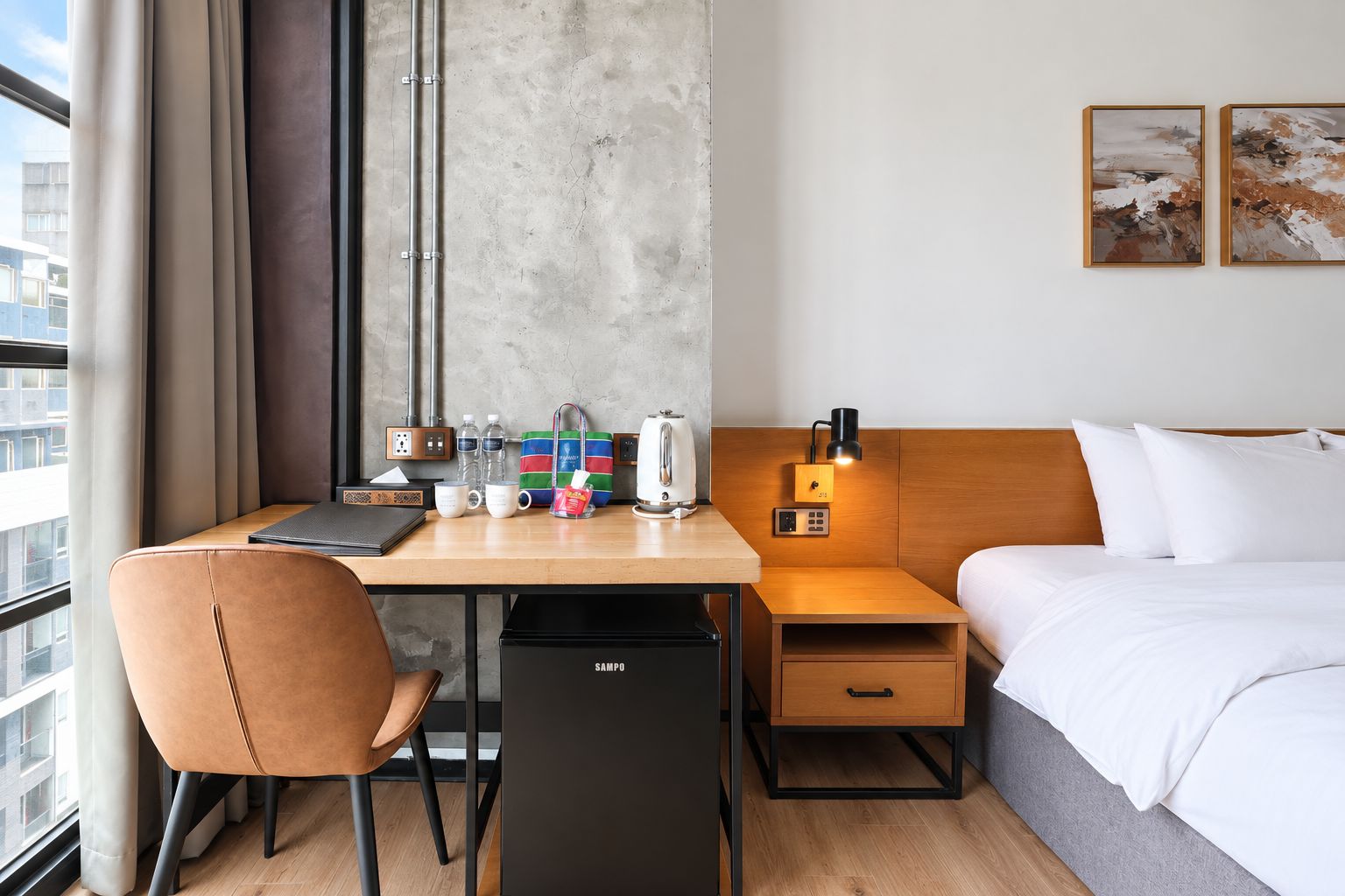 Una habitación de hotel con una cama, un escritorio con una silla, una mesita de noche, una lámpara y vistas de la ciudad por la ventana.