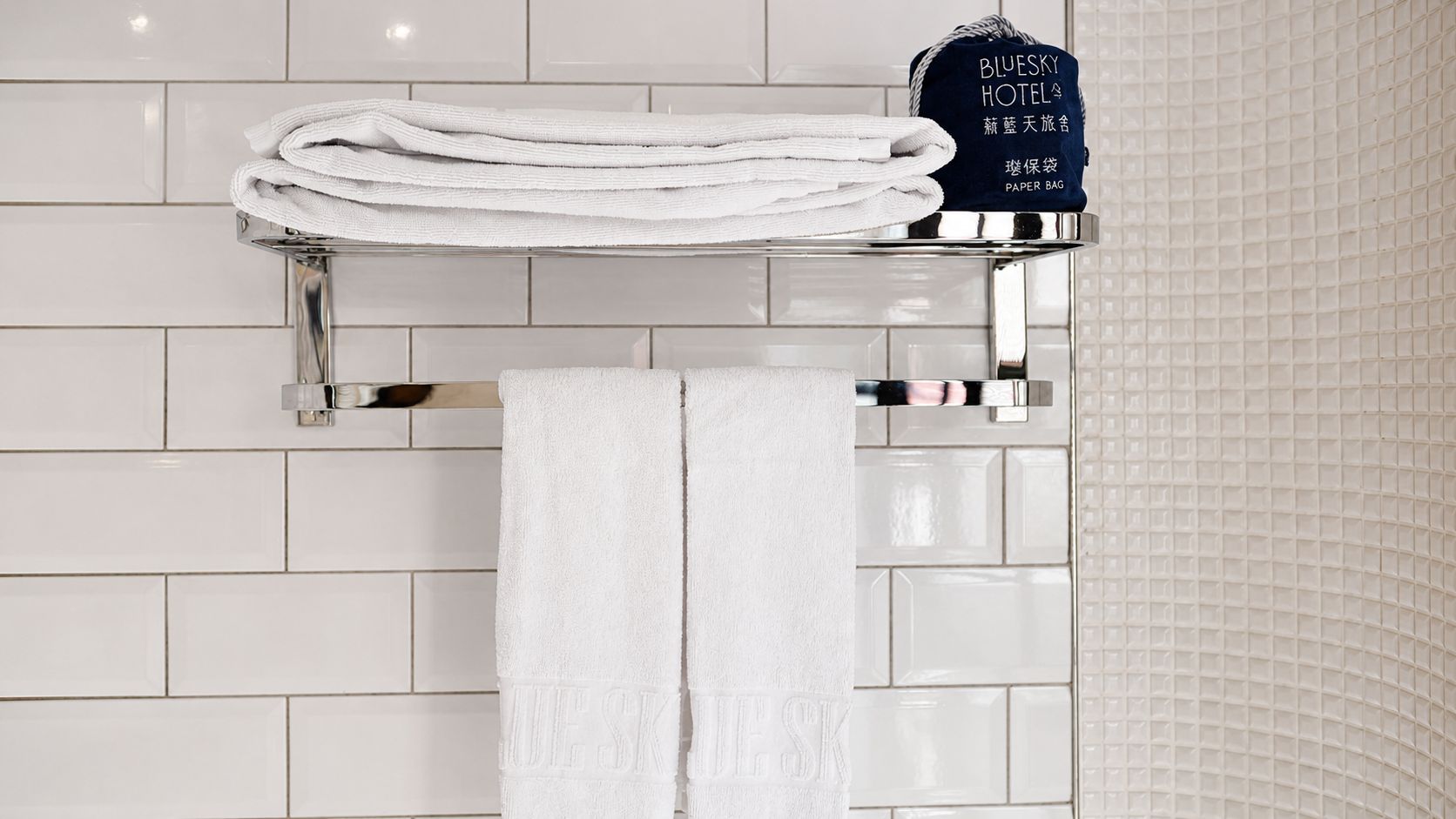 Un baño con paredes de azulejos blancos, un perchero con toallas dobladas y una bolsa de papel que dice 'Bluesky Hotel'.