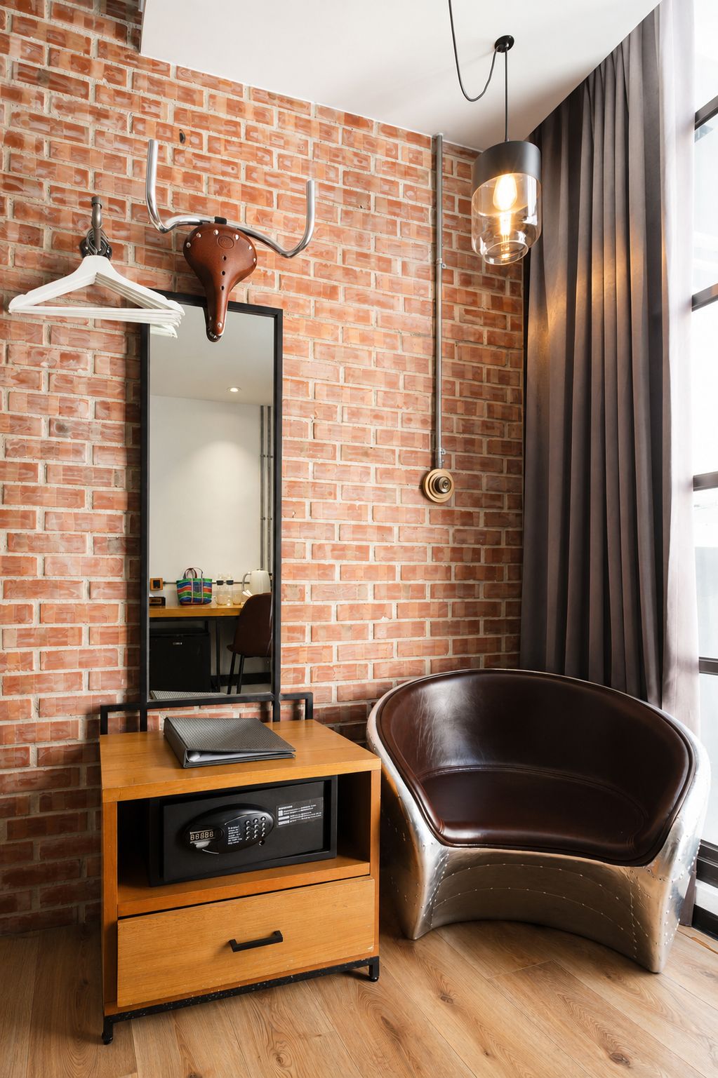 A room with a brick wall, a wooden desk, a leather chair, a mirror, and a hanging light fixture.