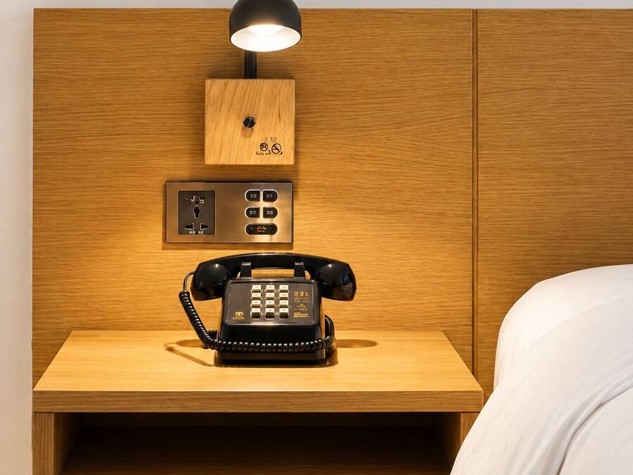 A wooden bedside table with a black telephone, a control panel, and a wall-mounted light fixture.