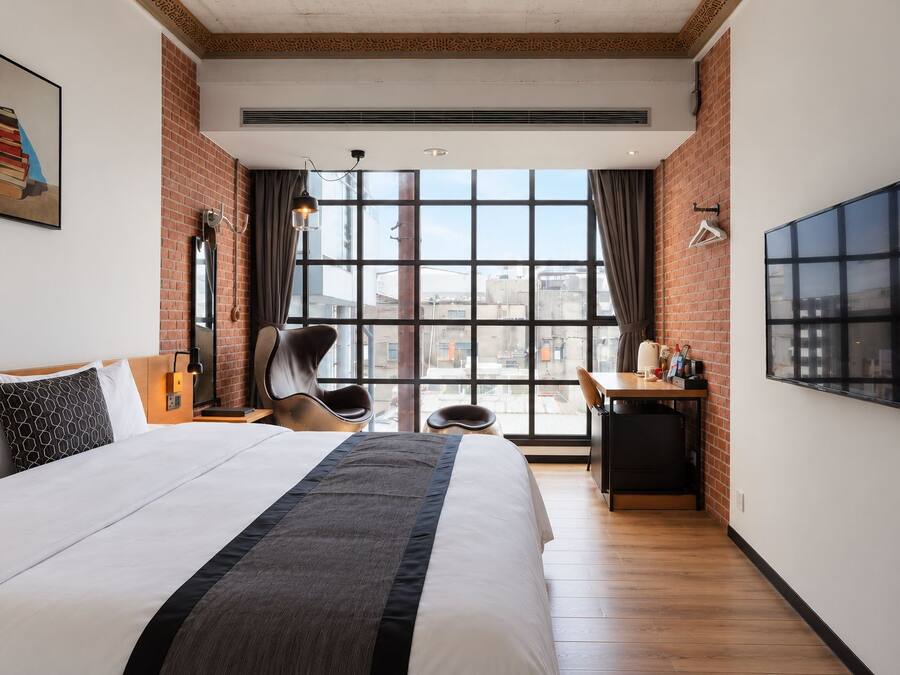 A hotel room with a large bed, a desk, a chair, a TV, and a window with a city view.