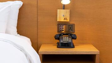 A neatly made bed with white linens, a wooden nightstand with a telephone and a lamp, and a wooden headboard.