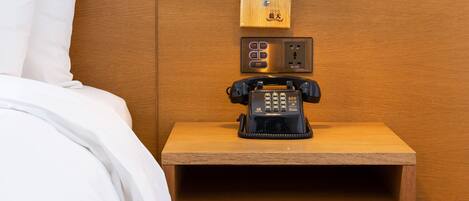 A neatly made bed with white linens, a wooden nightstand with a telephone and a lamp, and a wooden headboard.