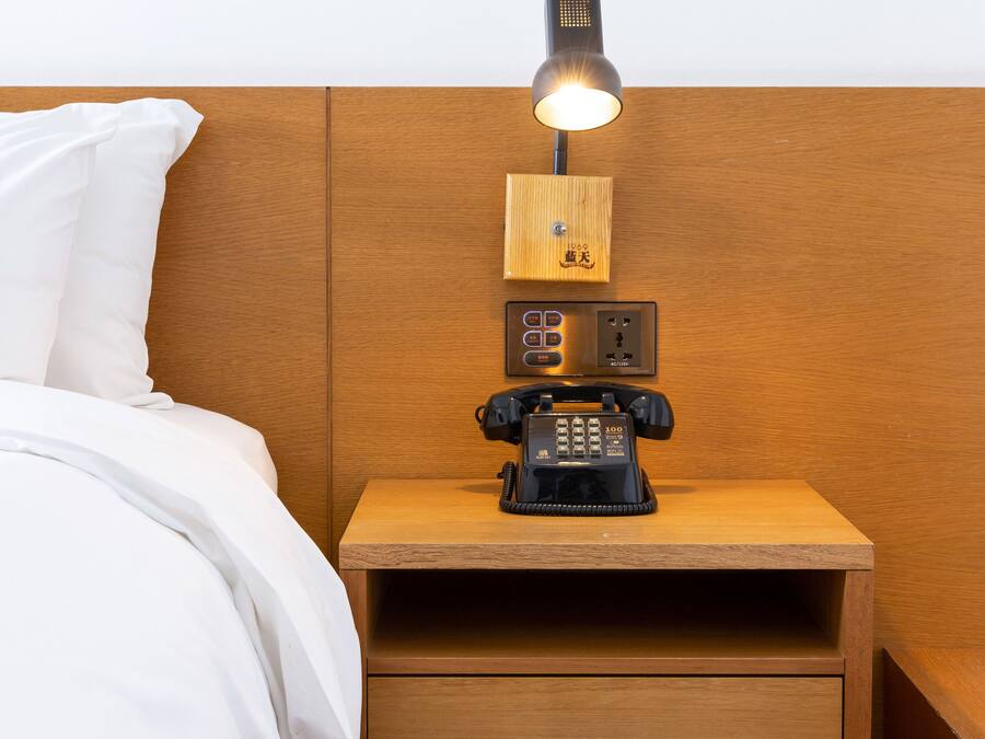 A neatly made bed with white linens, a wooden nightstand with a telephone and a lamp, and a wooden headboard.