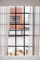 Habitación con un ventanal grande con diseño de cuadrícula, cortinas blancas y vistas a un edificio de ladrillo rojo con balcones.