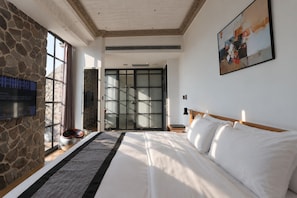 Habitación de hotel con una cama grande, una pared de piedra, un televisor y un ventanal corredero que da a un balcón.