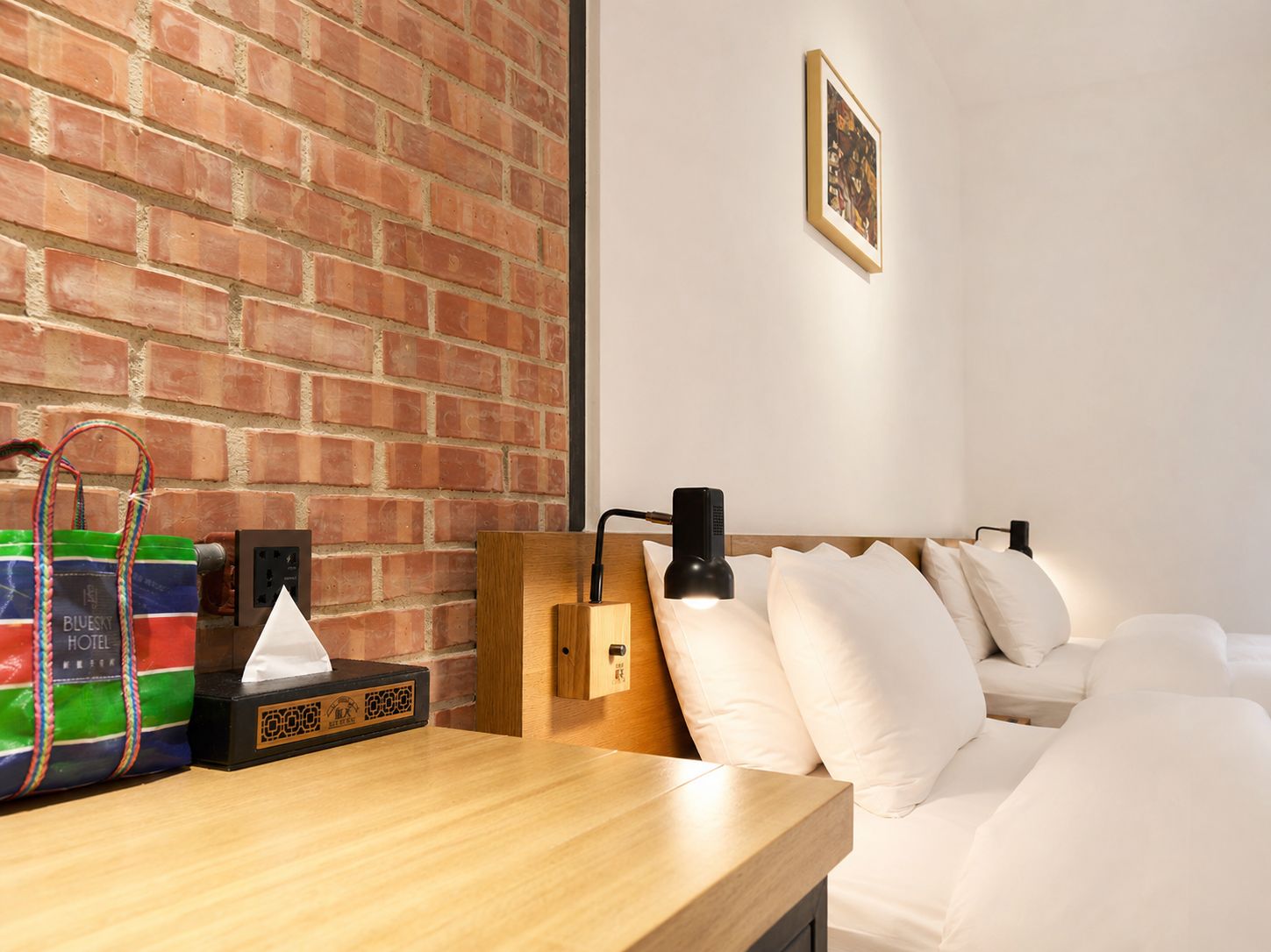 A hotel room with a brick wall, a wooden nightstand, a black lamp, a bed with white bedding, and a framed picture on the wall.