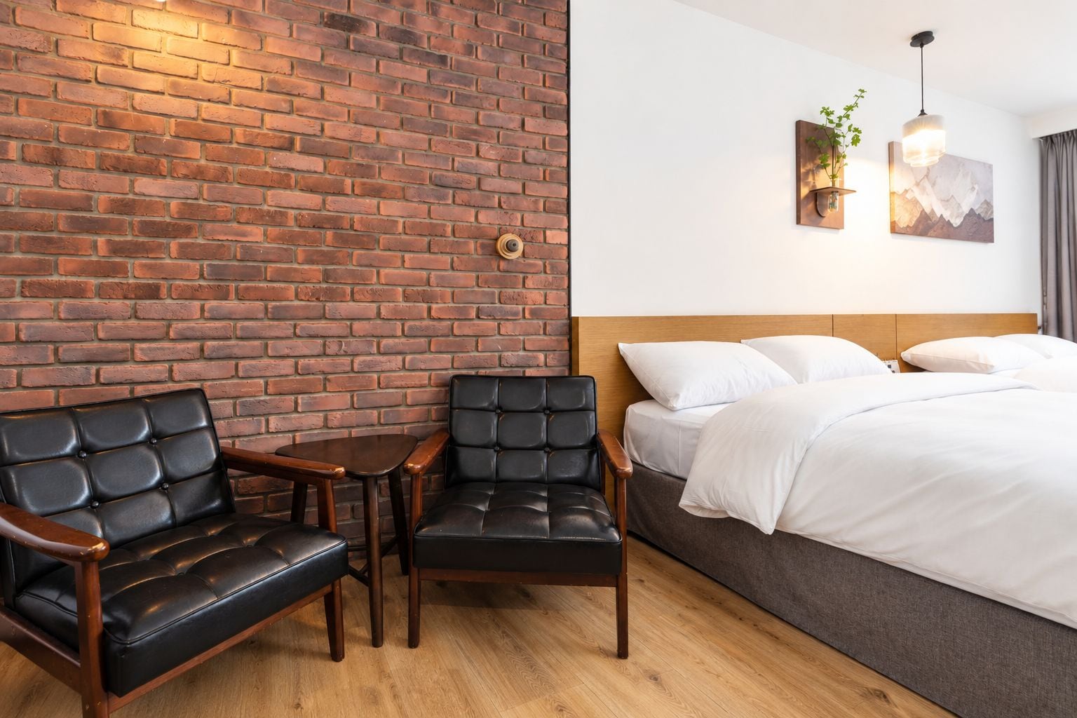 A hotel room with a brick wall, a bed with white bedding, two leather chairs, a small wooden table, and a wall-mounted light fixture.