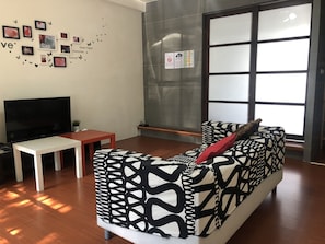A modern living room with a black and white patterned sofa, a flat-screen TV, a small white table, and a large window with blinds.