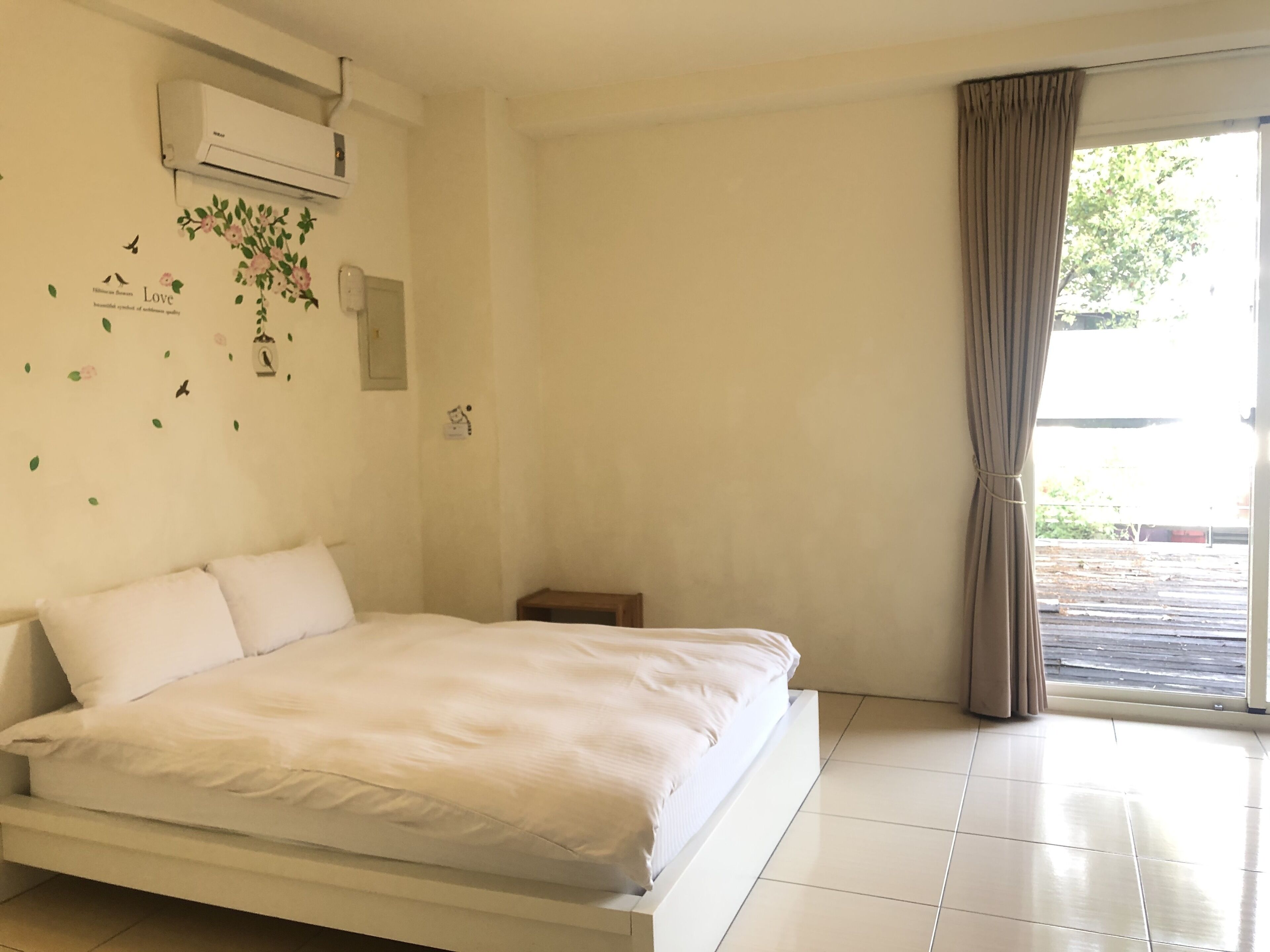 A bedroom with a bed, an air conditioning unit, a small bedside table, and a window with curtains.