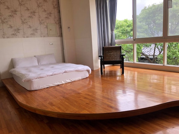 A bedroom with a curved wooden floor, a bed with white bedding, a chair, and a window with a view of trees.