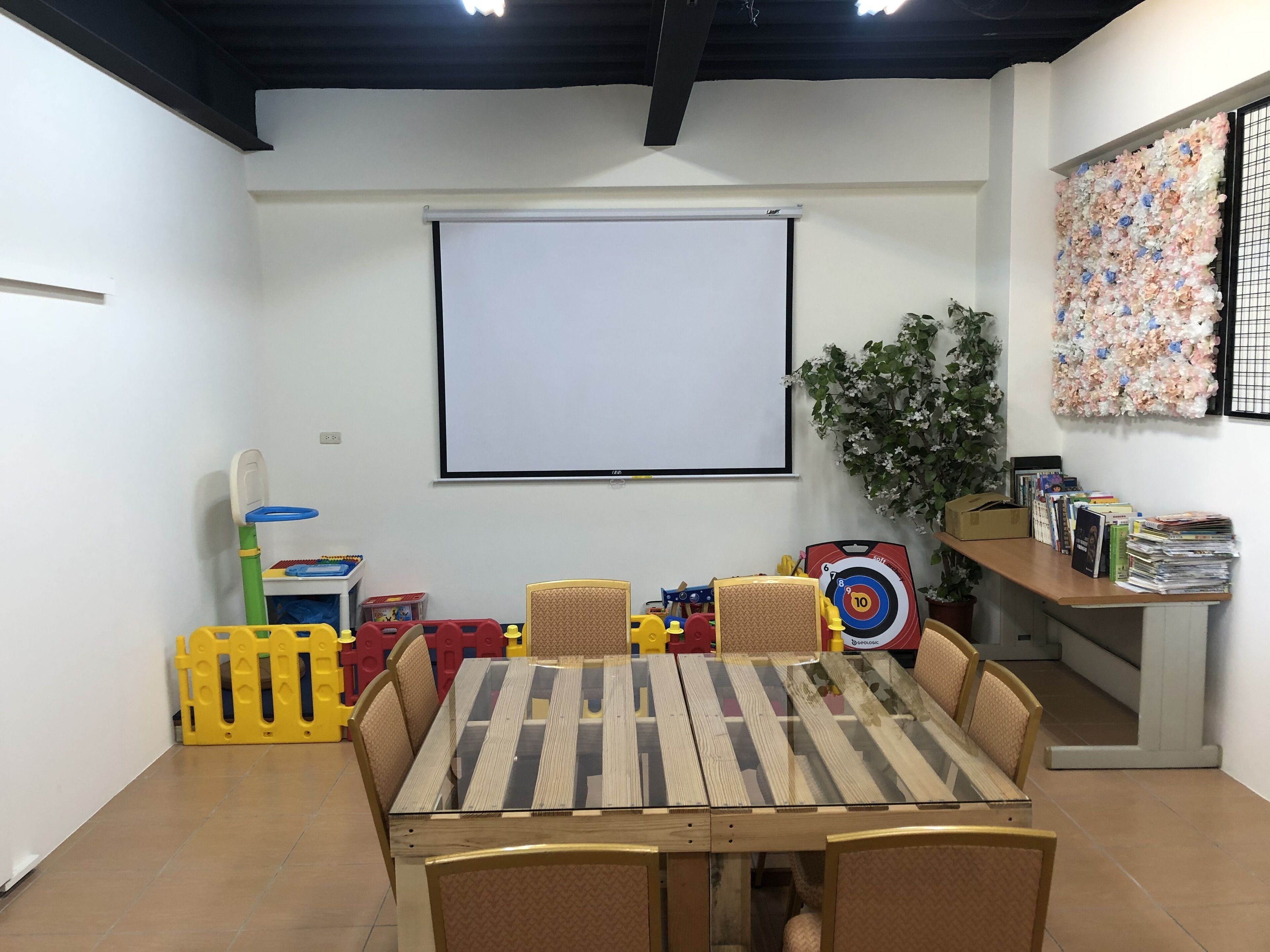 A room with a wooden table and chairs, a projector screen, toys, and a bookshelf.
