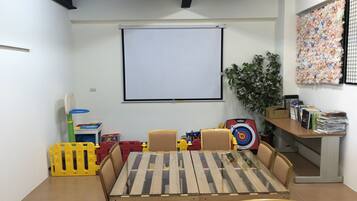 A room with a wooden table and chairs, a projector screen, toys, and a bookshelf.