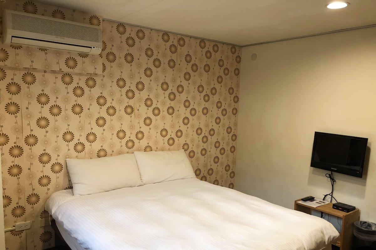 Une chambre d’hôtel avec un lit, un climatiseur, une télévision et une petite table de chevet en bois.