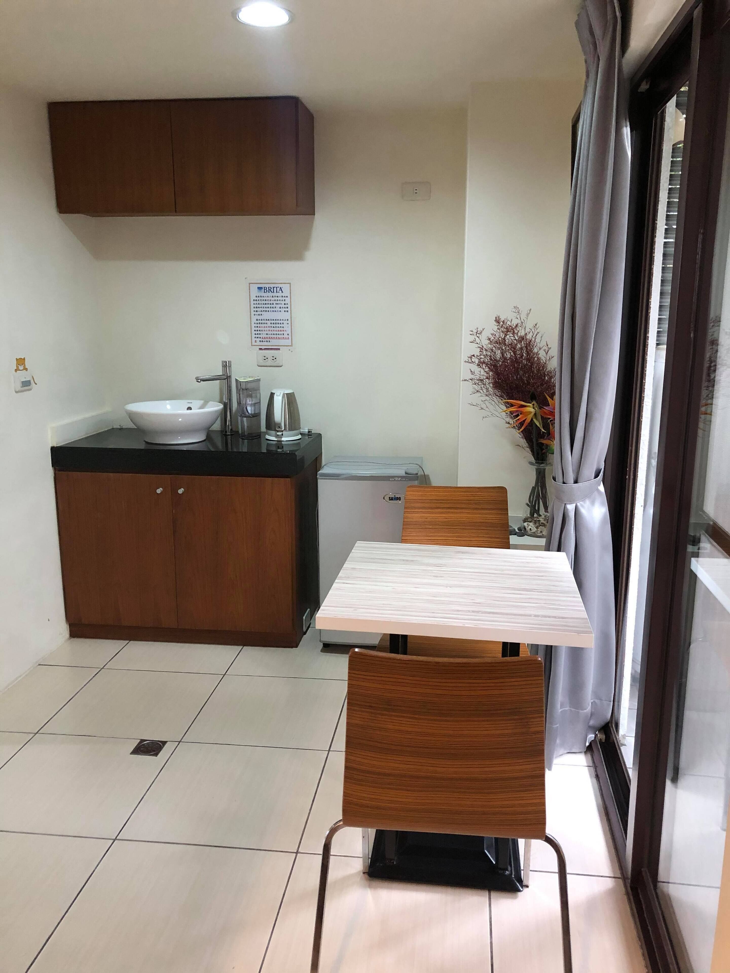 A compact kitchen with a sink, a coffee maker, and a small refrigerator. There is a dining table with chairs and a vase with flowers.