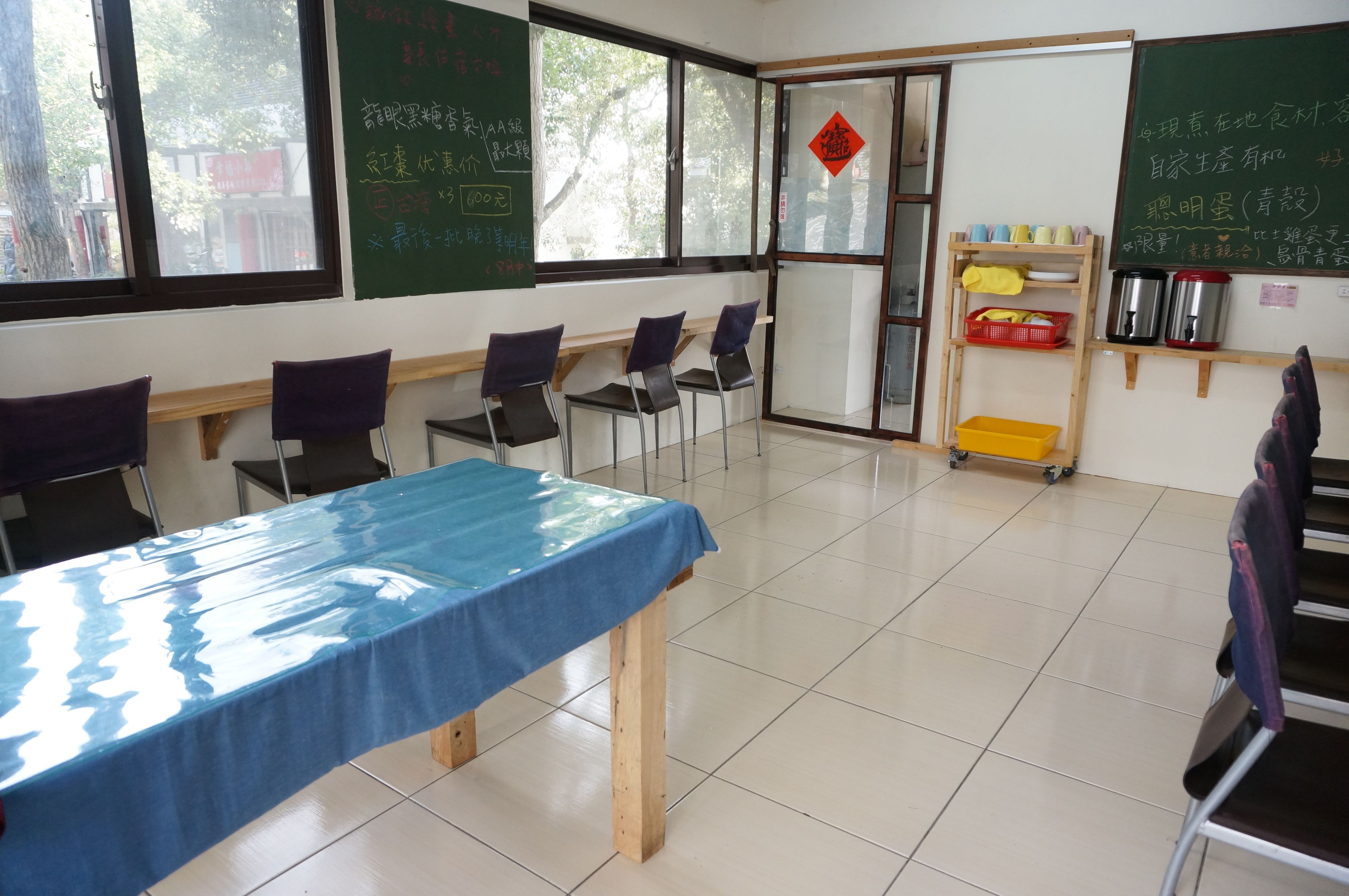 A classroom with a blue table, chairs, a chalkboard, and a window with a view of trees.